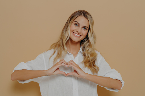Blonde young woman in white shirt making heart shape with her hands, displaying her love and admiration for someone while standing isolated in front of orange background. Appreciation concept