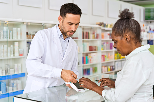 Senior woman paying by credit card in pharmacy, male pharmacists wearing lab coat standing behind the counter.