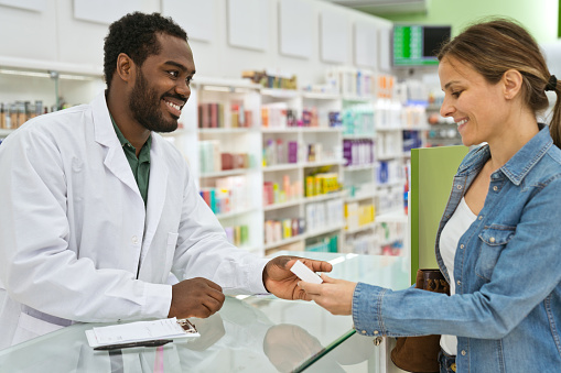 Male pharmacists talking with young woman in pharmacy, holding pills in hand.