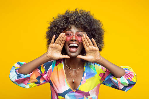 Happy young woman with afro hairstyle wearing colorful dress and pink eyeglasses shouting at camera. Studio shot on yellow background.