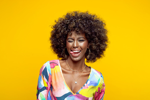 Happy young woman with afro hairstyle wearing colorful dress winking and sticking out tongue. Studio shot on yellow background.