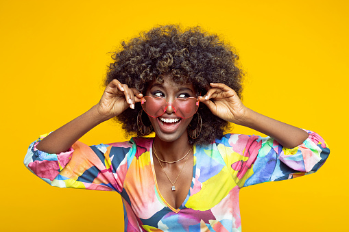 Excited young woman with afro hairstyle wearing colorful dress and pink eyeglasses looking away and laughing. Studio shot on yellow background.