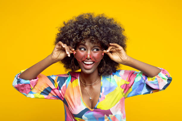 Summer portrait of beautiful young woman Excited young woman with afro hairstyle wearing colorful dress and pink eyeglasses looking away and laughing. Studio shot on yellow background. excited woman yellow stock pictures, royalty-free photos & images