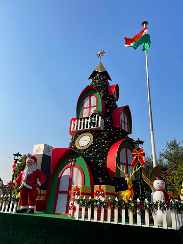 Stock image showing an Indian outdoor Christmas decoration, Father Christmas / Santa Claus is standing in front of a Christmas tree shaped house with presents, reindeer and snowmen on a sunny day, a blue sky in the background and a flag pole with the Indian flag.