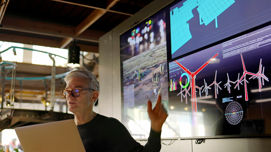 Stock photograph of a man conducting a seminar / lecture with the aid of a large screen. The screen is displaying data & designs concerning low carbon electricity production with solar panels & wind turbines. These are juxtaposed with images of conventional fossil fuel oil production.