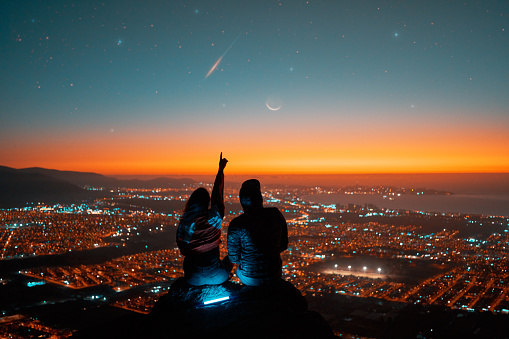 silhouette rear view of a heterosexual couple sitting on top of a mountain watching the starry sky with milky way and meteorite over the city