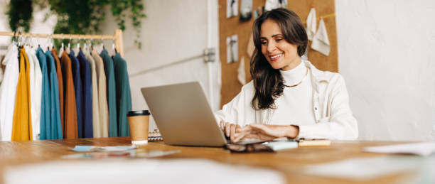 fashion designer happily working in her boutique, using a laptop to create her clothing designs - boutique owner store retail occupation imagens e fotografias de stock