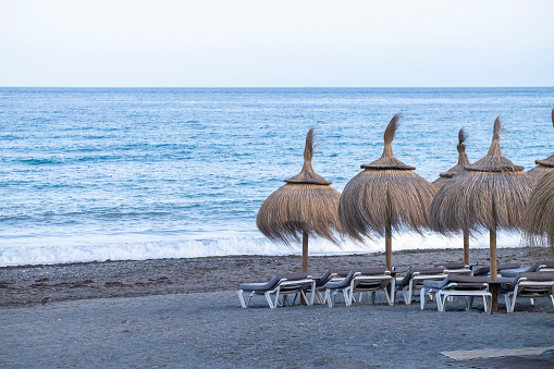 Straw beach umbrellas and comfortable sun loungers on clean sand and pebble beach. Costa del Sol. Mediterranean Sea. Vacation sea concept. Summer sunny seaside landscape.