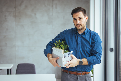 Unemployed guy in formal wear holding personal belongings, feeling depressed after losing his job. Upset Eastern man with cardboard box of things leaving office after being fired