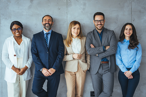 The business people standing on the gray wall background. Business team headed with boss, posing to camera over grey wall in office. Diverse businesspeople smiling at the camera