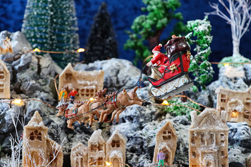 Stock photo showing a beautiful Christmas village display, which features numerous illuminated wooden houses. The houses have been placed on artificial grass and snow to create a snowy mountain scene, in front of a blue velvet backdrop, complete with a forest of plastic and wooden trees.