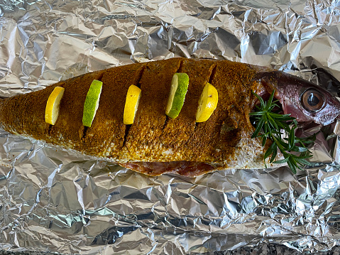 Stock photo showing a close-up, elevated view of fish being prepared to be cooked outdoors in a metal fish basket over the smoking flames of a portable, charcoal barbecue. Red snapper has been covered in a tikka spice dry rub and garnished with slices of lemon, lime and a sprig of herb.
