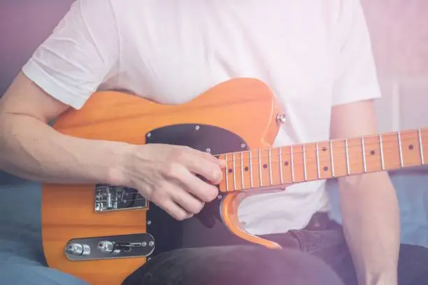 A man in a white T-shirt plays a natural-colored electric guitar sitting on a bed in close-up, selective focus.A male musician plays an electric guitar.Telecaster of natural color.Man playing guitar.