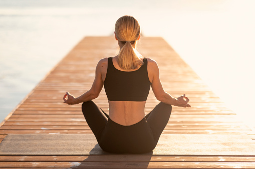 Morning Meditation. Rear view of sporty woman in activewear practicing yoga outdoors, unrecognizable athletic female sitting in lotus position on wooden pier near river, enjoying meditating