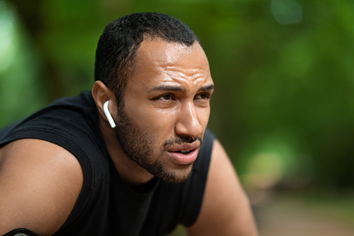 Closeup portrait of exhausted handsome african american young guy jogging in public park, having break while getting ready for marathon, listening to music, using earbuds, looking at copy space