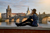 Stylish beautiful young woman wearing black hat sitting on Vltava river shore in Prague with Charles Bridge on background. Focus on background.