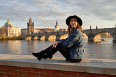 Stylish beautiful young woman wearing black hat sitting on Vltava river shore in Prague with Charles Bridge on background. Elegant retro lady fine art portrait.