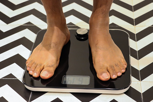 Stock photo showing close-up view of unrecognisable, Indian person, wearing grey bathrobe, standing on a pair of weighing scales in a luxury hotel bathroom with black and white zig-zag patterned floor and wall tiles. Dieting and eating disorder concept.