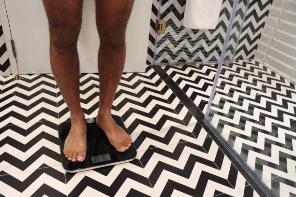 imagen de una persona irreconocible con los pies descalzos de pie sobre básculas, cabina de ducha de vidrio con paneles y puerta de baño blanca, baño de hotel de lujo monocromo en zig-zag con paneles de pared y baldosas con motivos de piso, enfoque en p - barefoot behavior toned image close up fotografías e imágenes de stock