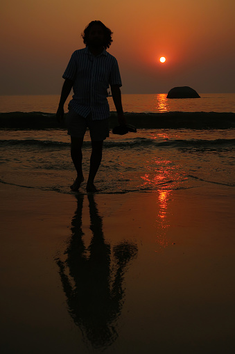 Stock photo showing an Indian man on holiday in Goa, South India, pictured walking on Palolem Beach, paddling in the gentle sea waves at sunset, a particularly popular winter holiday destination for both English and German tourists.