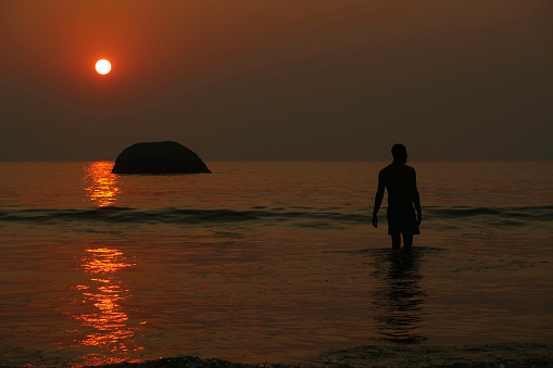 Stock photo showing an Indian man on holiday in Goa, South India, pictured walking on Palolem Beach, paddling in the gentle sea waves at sunset, a particularly popular winter holiday destination for both English and German tourists.