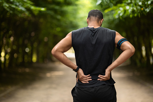 Back view of african american sportsman touching sore zone on his back, having back pain during exercising at public park, panorama with empty space. Workout injuries concept