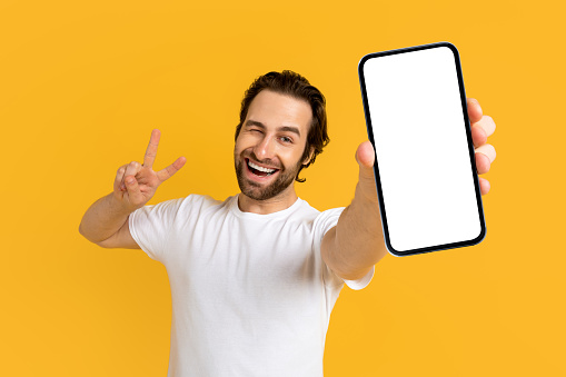 Cheerful emotional young european guy in white t-shirt show phone with empty screen and peace sign with hand isolated on yellow background, studio. Gesture, big sale, new app, device, app and offer