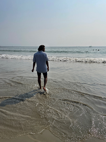 Stock photo showing an Indian man on holiday in Goa, South India, pictured walking on Palolem Beach, paddling in the gentle sea waves, a particularly popular winter holiday destination for both English and German tourists.