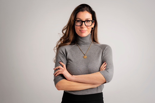 Studio portrait of an attractive middle aged woman with eyewear wearing turtleneck sweater and black pants while standing at isolated grey background. Copy space. Studio shot.