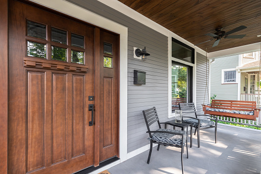 A wooden front door on a grey home's covered front porch with chairs and a wooden hanging swing.