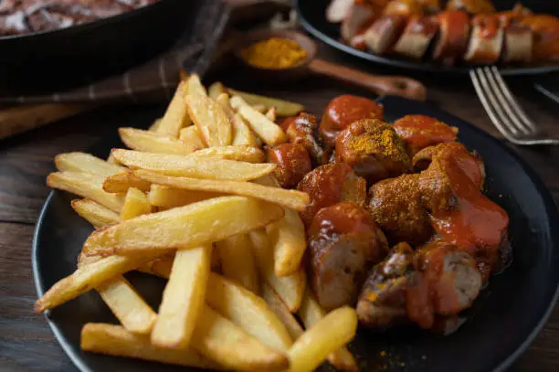 Delicious german fast food meal with homemade french fries and traditional curry sausage with delicious curry sauce. Served on a dark plate on rustic and wooden table background. Closeup