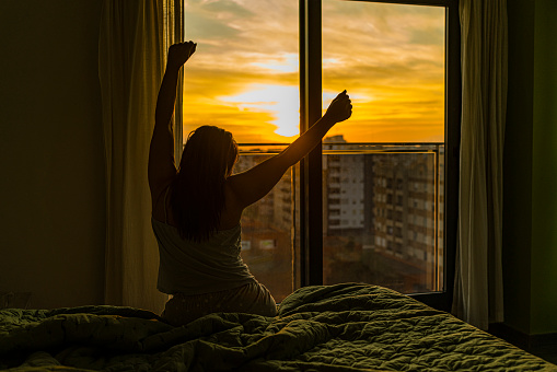 Rear view of unrecognizable woman sitting on bed in the morning, looking through window and stretching arms. High resolution 42Mp indoors digital capture taken with SONY A7rII and Zeiss Batis 40mm F2.0 CF lens