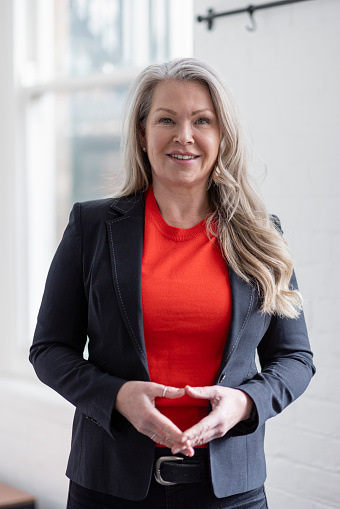 A close-up waist-up shot of a mature confident businesswoman looking into the camera with her hands together. She is dressed smartly in a suit jacket and standing in an office interior. She is based in the North East of England.