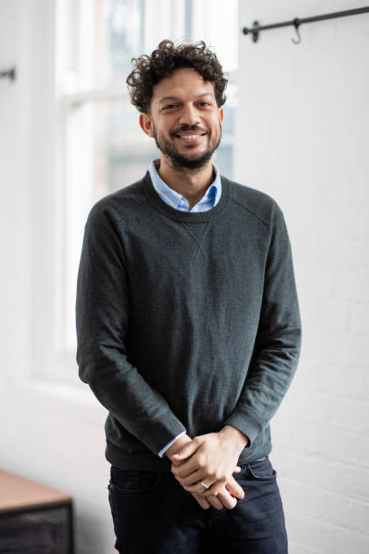 A close-up three quarter length shot of a mid adult confident businessman looking into the camera with his hands together. He is dressed smartly and standing in an office interior. He is based in the North East of England.