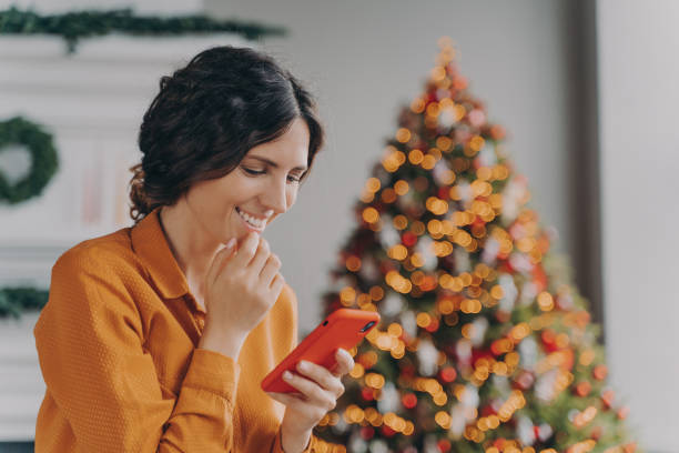 istock Overjoyed hispanic lady sits in living room decorated with Xmas tree looking at mobile screen 1475065652