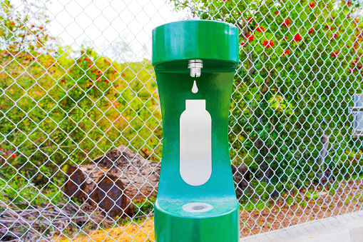 Green water bottle filling station set against a mesh wire fence outdoors. Convenient access to clean drinking water while hiking.