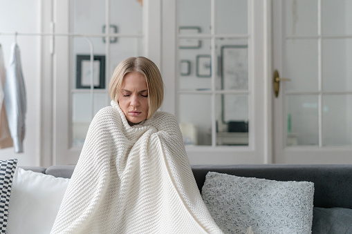female feeling sick and has influenza wrapped with knitted soft plaid and sitting on sofa in living room, girl with temperature and grippe alone at home