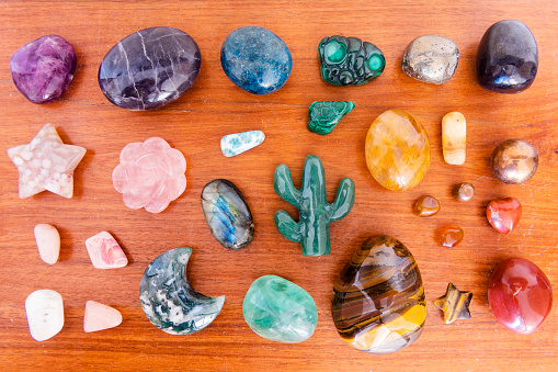 This is a color photograph of an assorment of both tumbled and carved crystals ranging from red jasper, orange carnelian, tiger’s eye, yellow arragonite, pyrite, copper, shungite, malachite, aventurine, green and purple flourite, labradorite, moss agate, flower agate, rhodochrisite, iolite, blue apatite, larimar, golden healer, rose quartz, and scolicite.