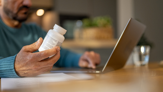 Mature man holding pill bottle while using laptop in kitchen.