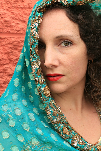 A closeup of a Caucasian woman with a green shawl over half her face. She is wearing medium length brown curly hair , makeup and a necklace.