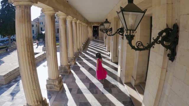 Aerial shot of tourist woman in red dress and hat walking exploring old european city narrow streets on Corfu, Greece island sightseeing local architecture, slow motion