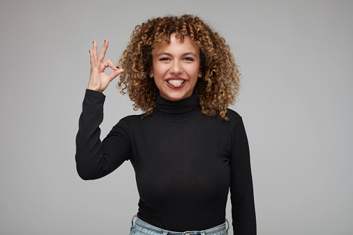 Cute young woman with curly hair sticking out her tongue and making a hand gesture saying everything is fine.