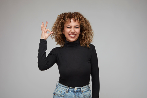 Cute young woman with curly hair sticking out her tongue and making a hand gesture saying everything is fine.