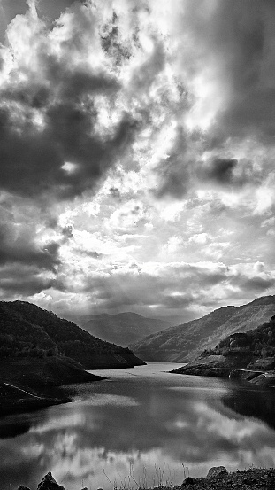 A black and white vertical shot of a Valley in north West Turkey. There is a lake in between the hills on both sides and in the distance at the back, the water curves on the sides of the hills like a giant snake. The sky is cloudy with the sun shining behind them and this is reflected on the surface of the water.