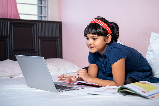 Portrait of smiling little girl using laptop while study online at home and looking at camera