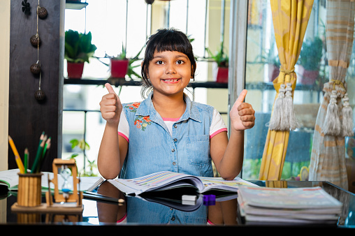 Portrait of an adorable little girl posing with her thumbs up while doing her homework at home