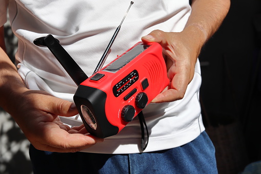 A man holds in his hands a small red portable radio rechargeable with solar panels or manually with a crank and an included flashlight