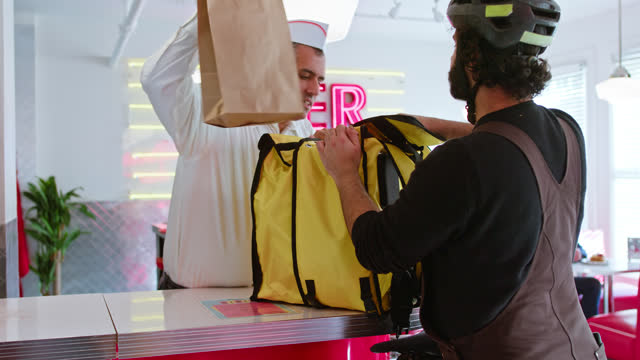 Man In a White Button Down Shirt Puts Order Inside Delivery Person's Bag In a 1950s Styled Diner