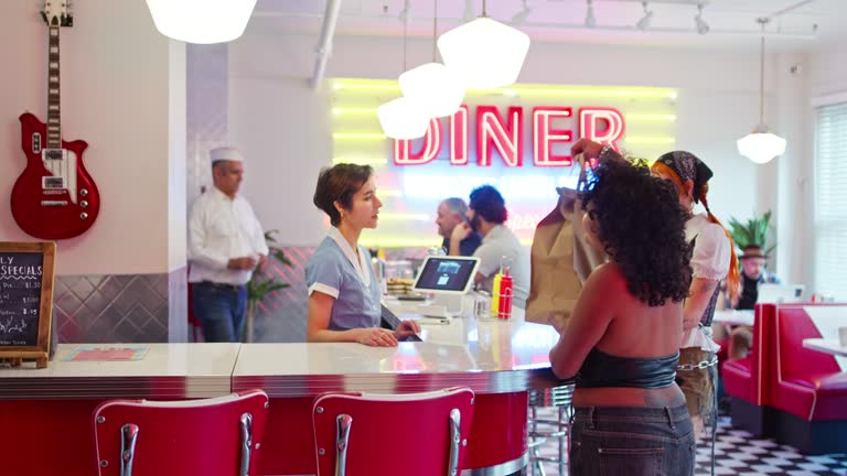 Shot Of Waitress Handing Order To Two Non-Binary People In 1950s Styled Diner