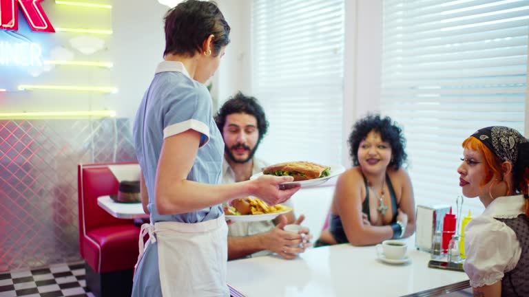 Slow Motion Shot Of Waitress Serving Food To Two Non-Binary People And a Man In 1950s Styled Diner